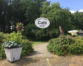 Eingangsschild des Cafés mit Blumen und Bäumen im Hintergrund.